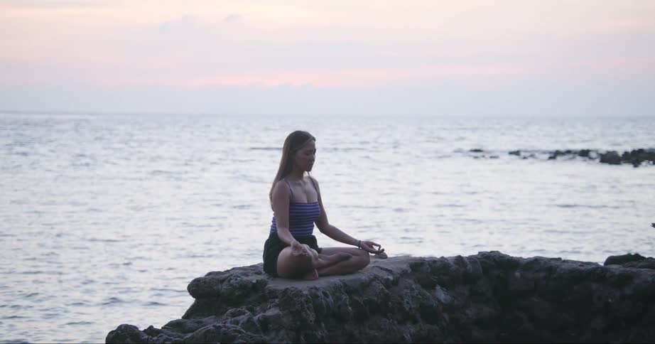A woman meditates while sitting on a cliff over the ocean