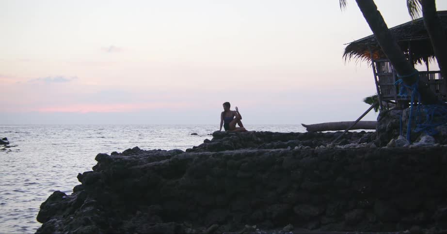 A woman is doing a yoga pose on a rock overlooking the ocean at dusk