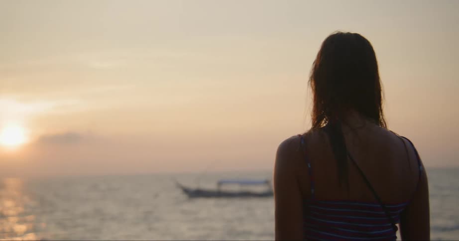 Woman with brown hair and swimsuit watches boat and sea at sunset