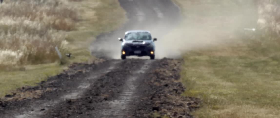 A vehicle driving on a dirt road surrounded by grass with a bird flying away