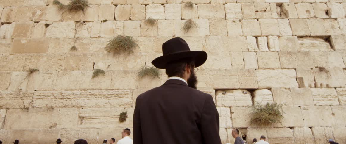 A bearded man stands in front of a stone wall