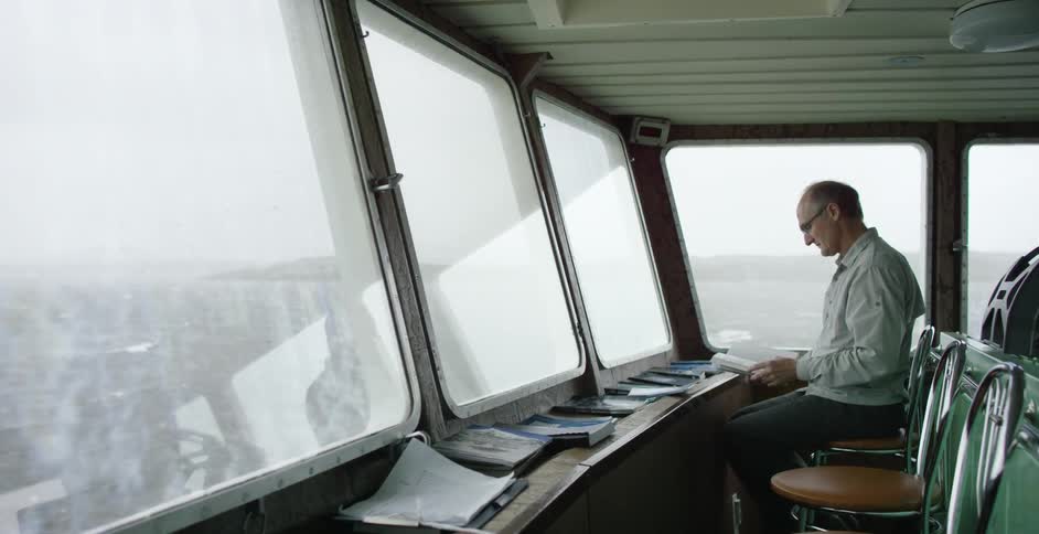 A man is sitting by a window looking over the water reading a book