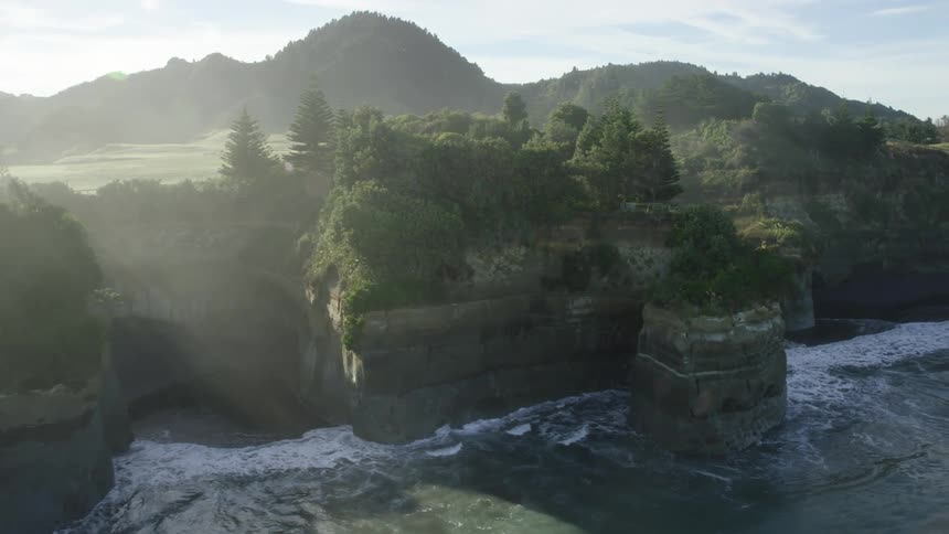 Waves crash against cliffs topped by woods that stand across fields from forest covered mountains