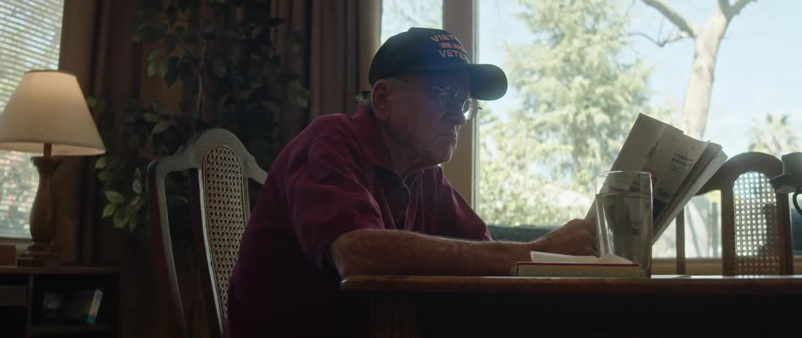 An older man wearing a veterans cap sitting at his dining room table reading a newspaper