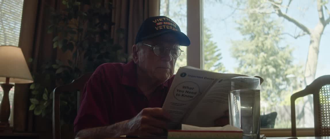 Elderly man with glasses dark blue cap and crimson shirt reads newspaper
