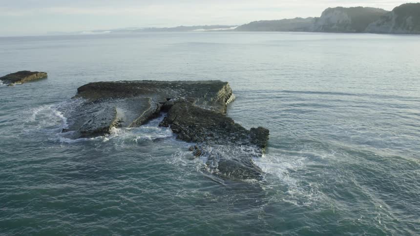 Wide open views in a drone shot of the ocean crashing against rocks and coastline with cliffs in the distance