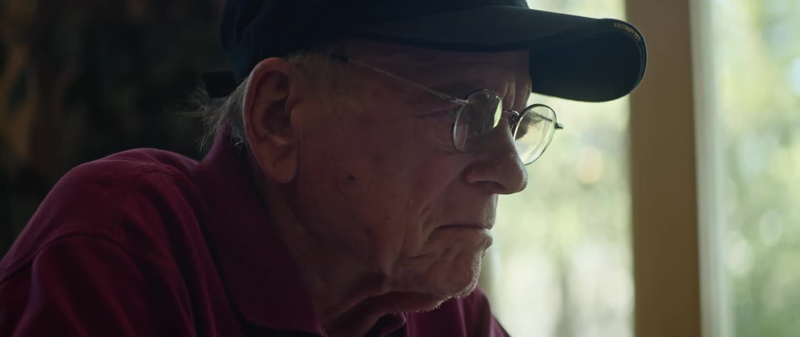 A man wearing glasses and a cap is sitting near a window as he shakes his head