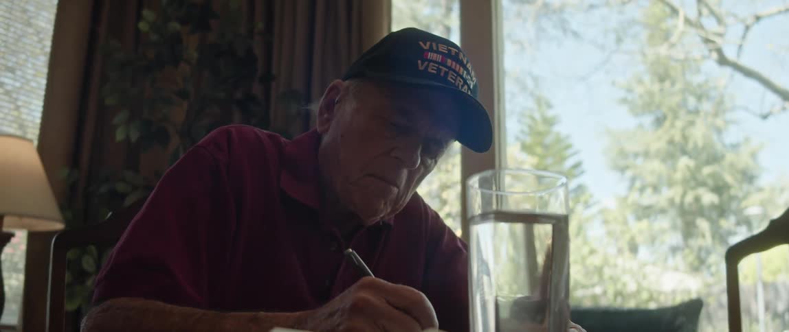 A man is sitting at a table with a glass of water next to a window as he writes on paper