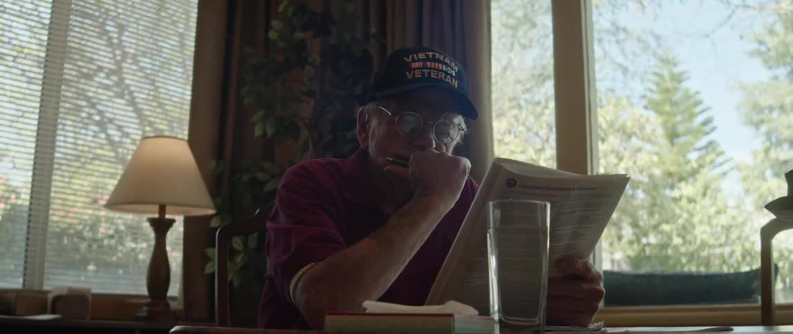 A man is sitting at a table in a restaurant next to a window with a glass of water as he reads a magazine