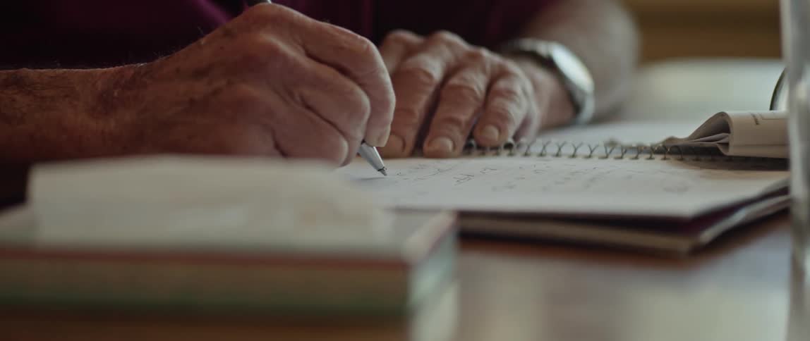 A man is sitting at a table writing in a spiral notebook