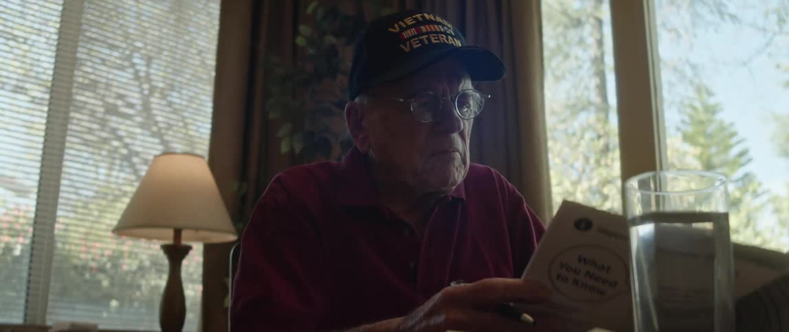 An elderly man sits indoors while reviewing a printed document