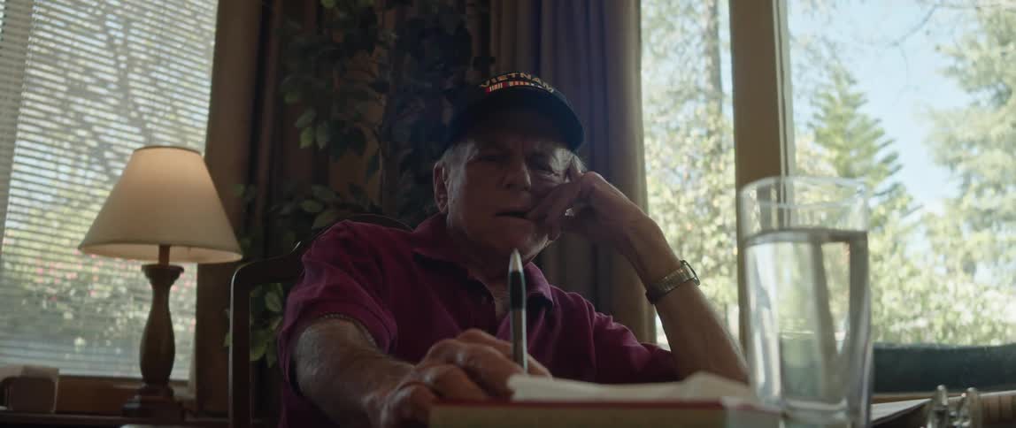 An older man sitting at his dining room table tapping a pen on the surface looking contemplatively