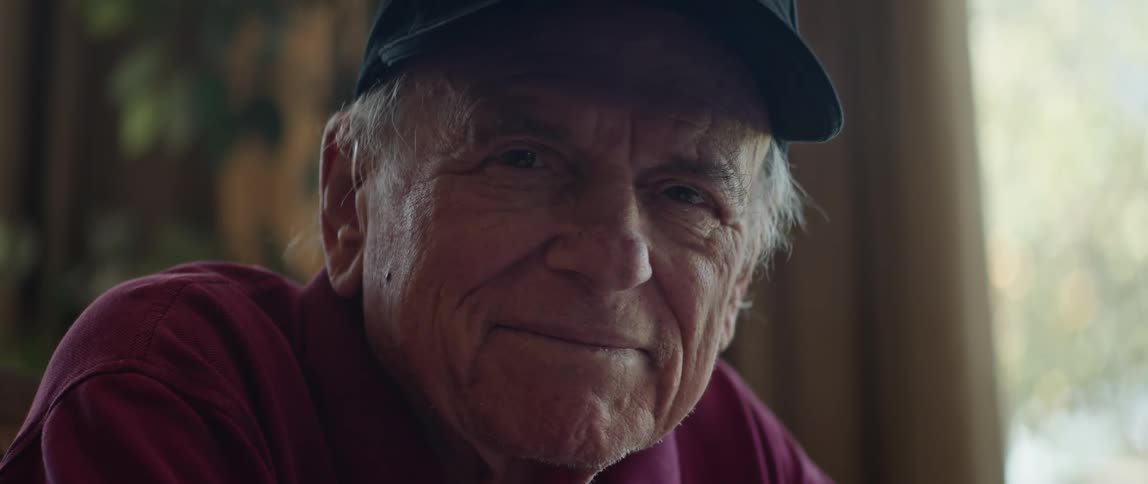 Elderly man with maroon shirt and black cap smiles at camera