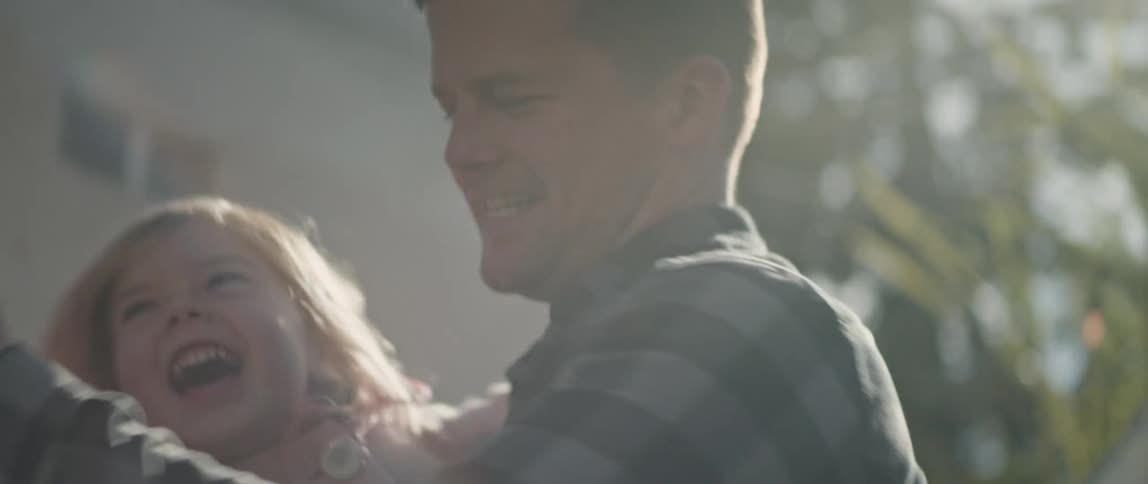 A man is standing outside in his yard near his house as he holds his little girl and spins her around as they laugh