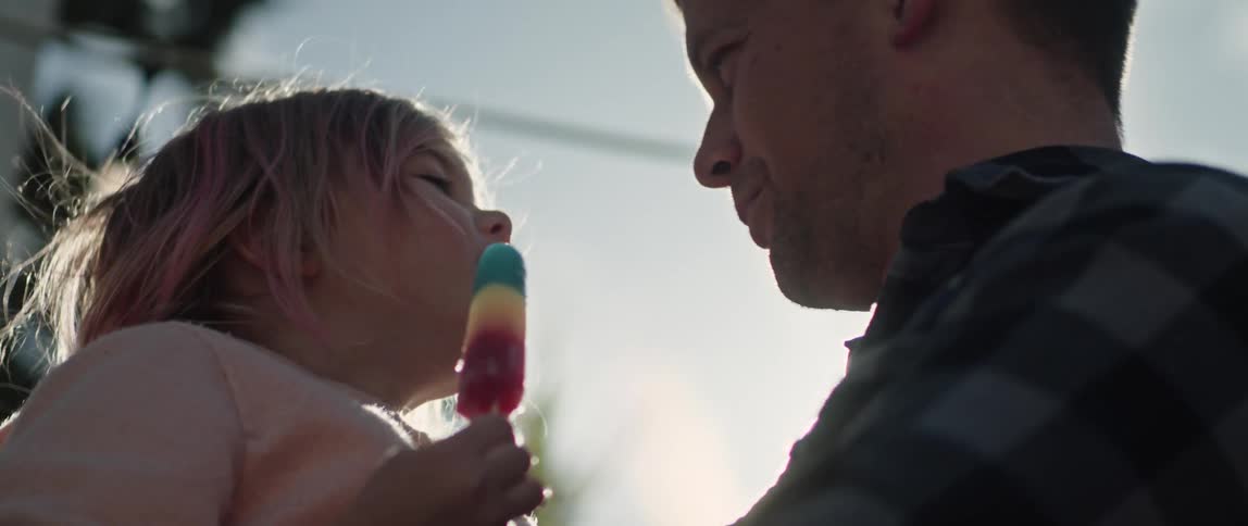 A father holds his daughter while she kisses him on the nose and he kisses her back while she holds a popsicle
