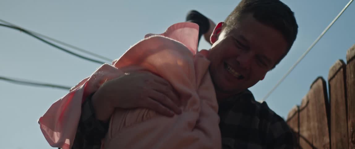 A man is standing outside in his yard near a fence with his little girl laughing on his shoulders and he flips her upside down with her bare foot in front of his face