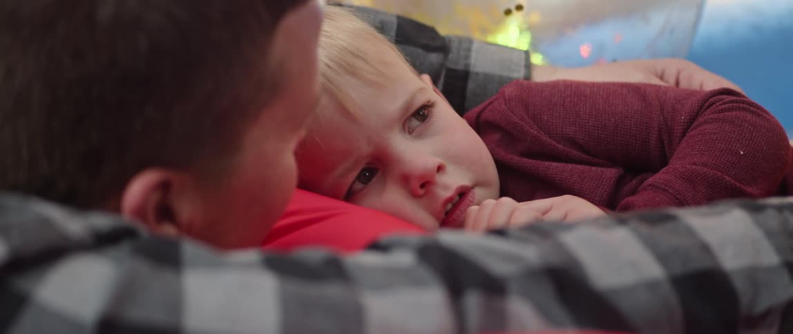 A little boy is lying on his fathers chest as he looks up and smiles and puts his fingers in front of his mouth