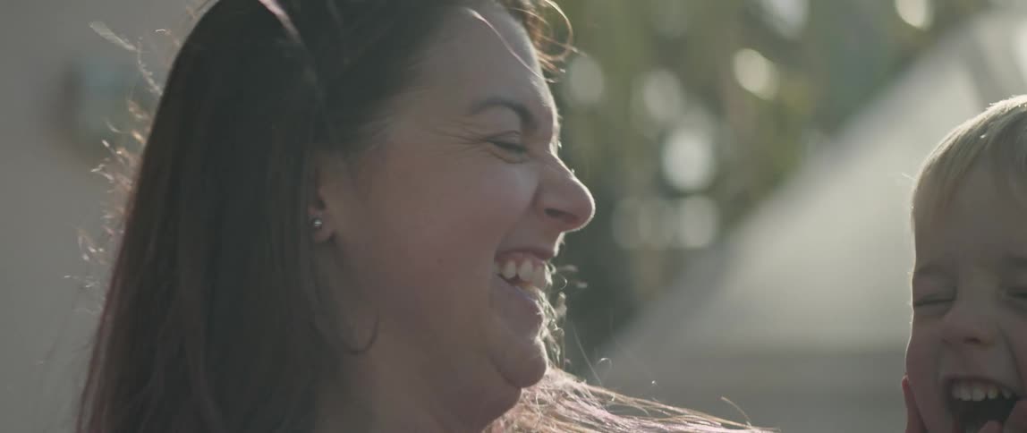 A woman is sitting outside holding her little boy and they kiss and laugh