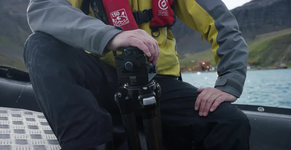 A person sitting on a boat holds onto a camera tripod