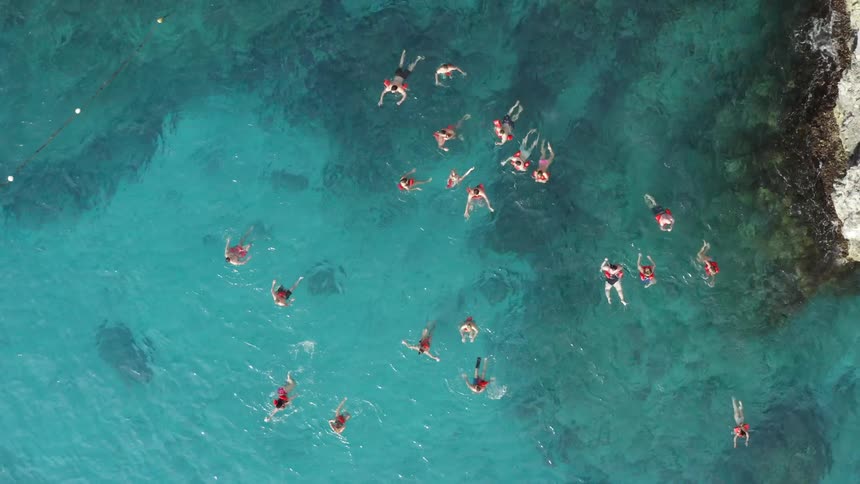 Aerial view of a dozen or more swimmers in beautiful blue caribbean water near rocky shore