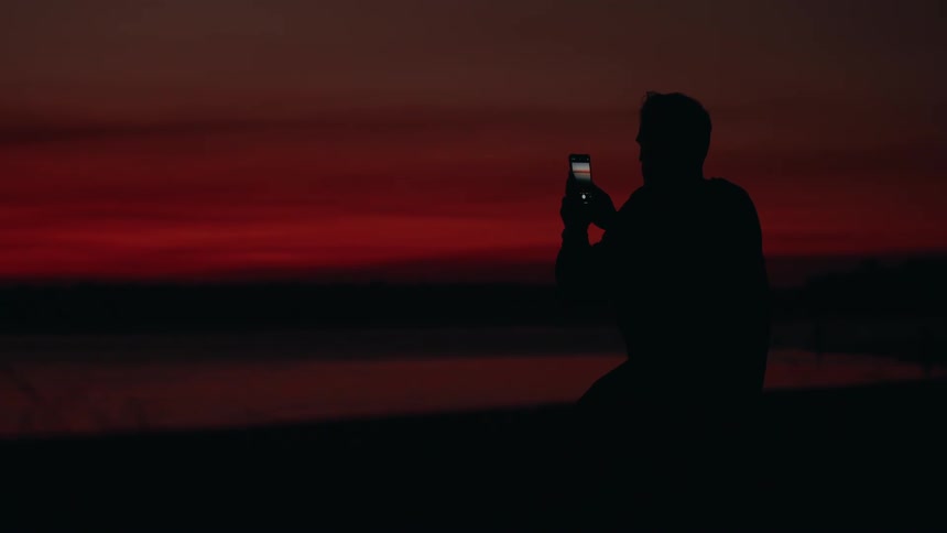 A silhouette of a man sitting outside using his cellphone to take a picture of the sky at dusk