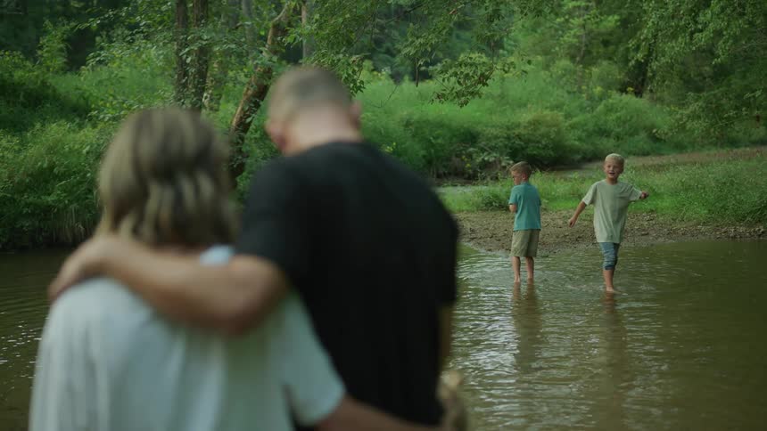 Mother and father stand on shore watching their kids wade in stream through wilderness