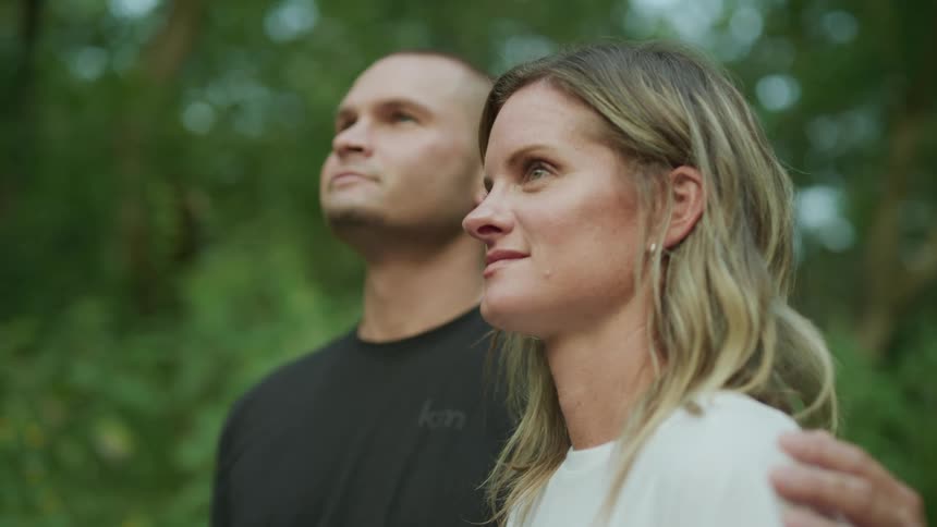 A man and woman standing next to each other the man with his arm around the womans shoulder as she looks down and smiles