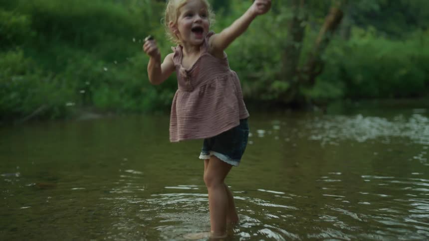 Blonde girl with shirt and shorts plays in the river next to green forest