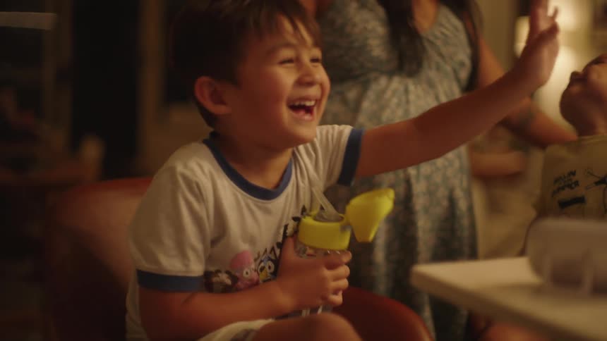 A child holding a water bottle laughs as his mom and brother are in the background