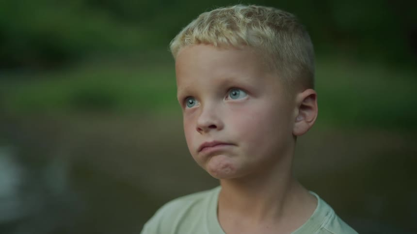 A young boy standing looking like he is thinking outdoors with kids playing behind him