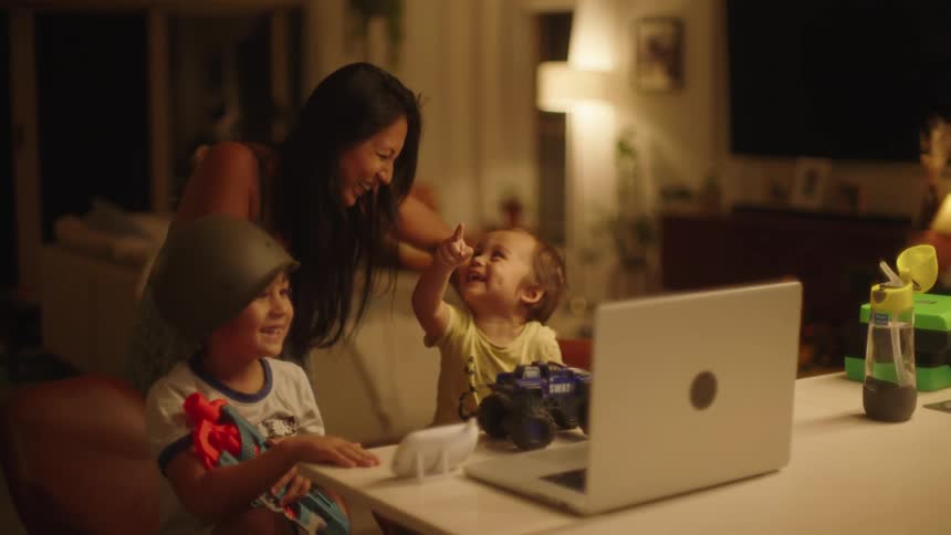 Mother stands behind her kids and laughs as they watch laptop
