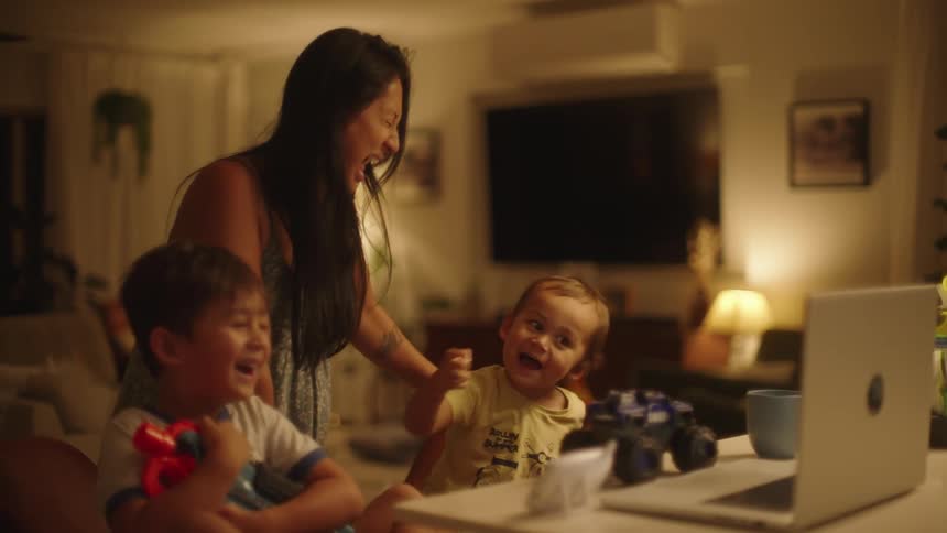 Two children are sitting at a table looking at a laptop computer and laughing as their mother stands behind them