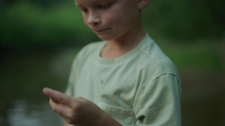Blonde boy with green t shirt examines pebble in the river next to forest