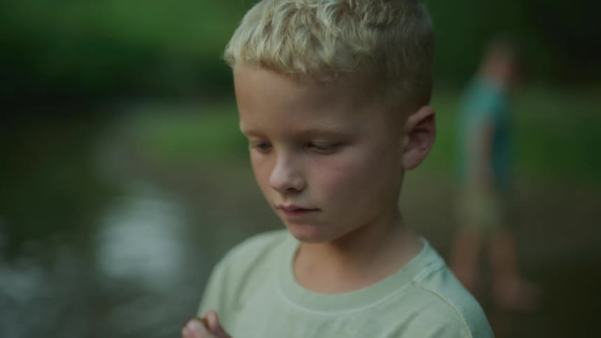 A boy standing looking at a pebble in his hand contemplatively