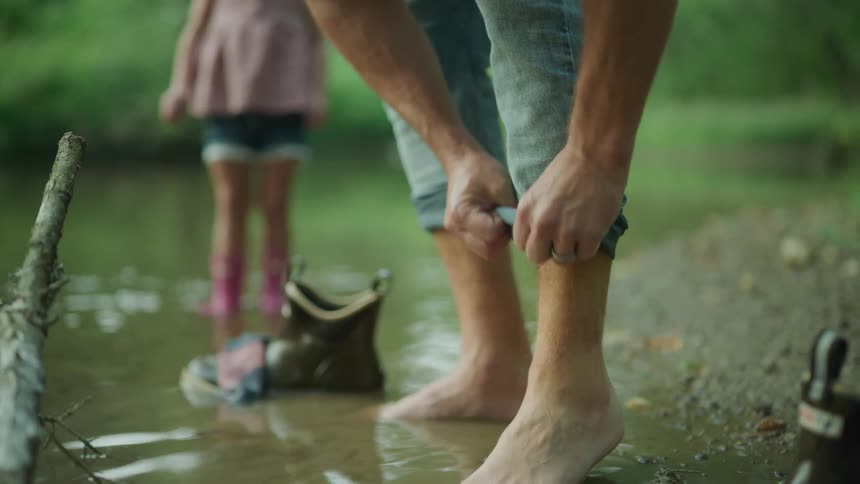 A woman rolls up her pants while standing on the riverbank with toes in the water