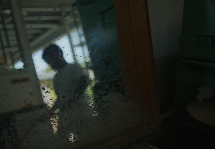 Young boy sweeping a room at home seen through a dirty window