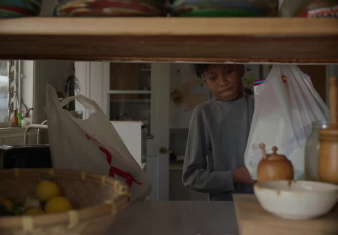 Two boys walk into a house and one boy puts grocery bags on the kitchen counter and takes things out of a bag