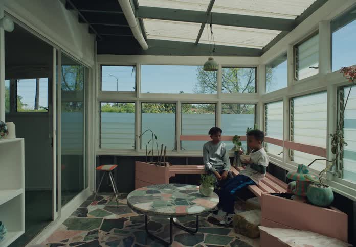 Two boys sit in a sun room on and one of them eats from a bowl