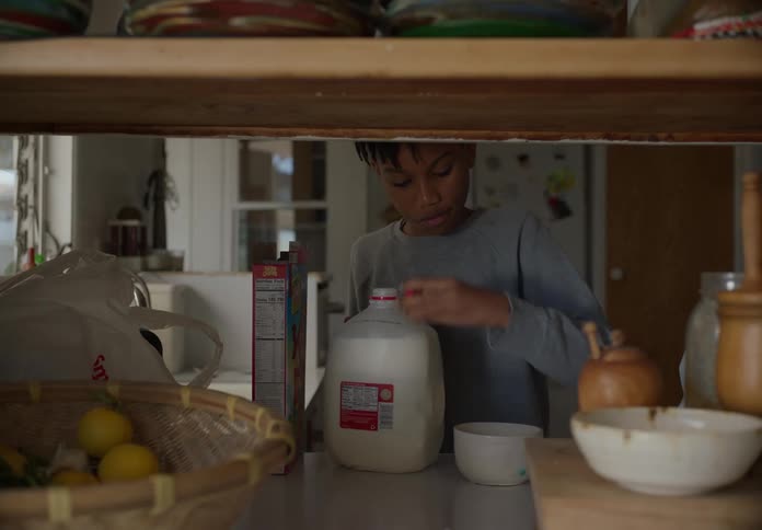 A boy pours himself some cereal and milk in a bowl while in the kitchen