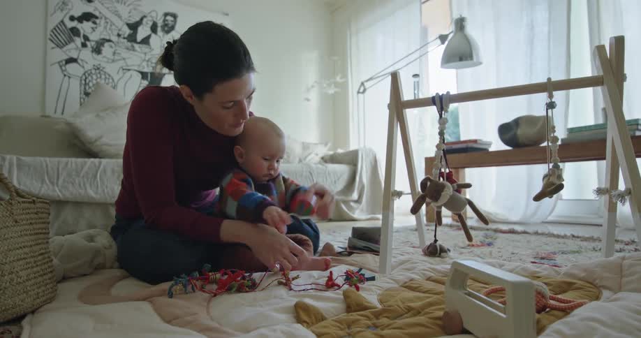 A woman is sitting on the living room floor paying with her baby and there are toys all over the floor