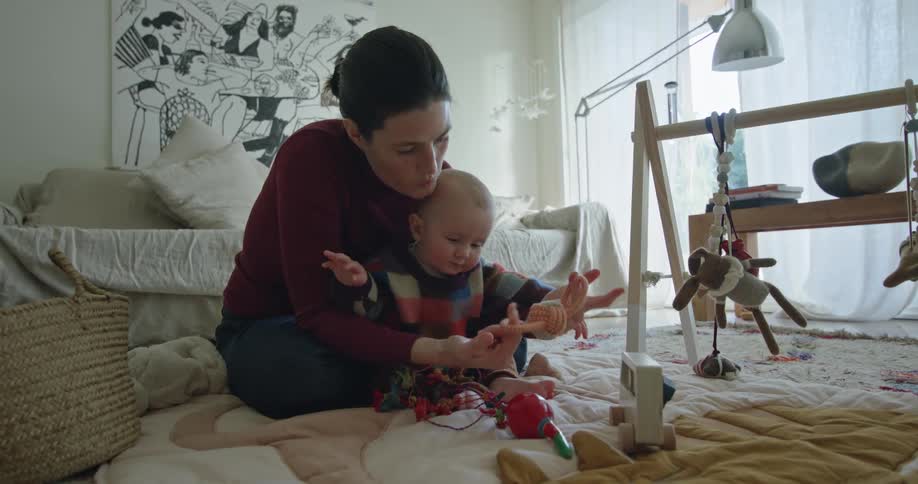 Amother sits on the floor with her baby while it plays with toys