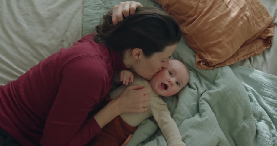 A mother kisses her baby daughters face while they lay on the bed