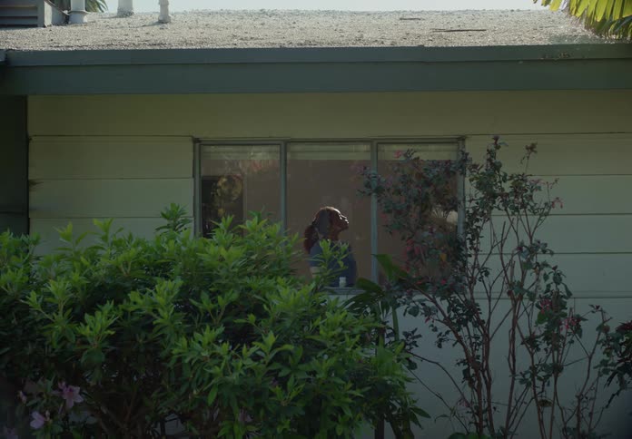 A teenage girl stands in the picture window of her house and longingly gazes up at something as if mesmerized