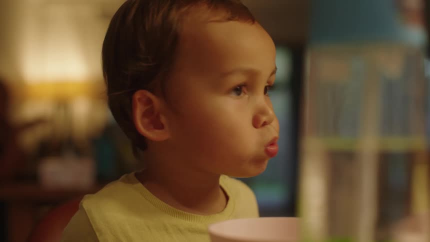 Toddler boy sitting at dinner table eating with a mouthful of food looking around wideeyed