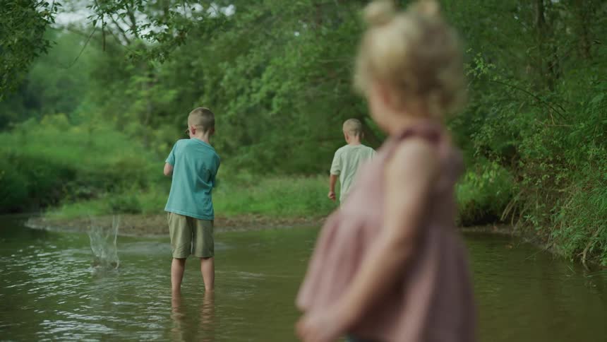 Two young boys and their little sister standing in the lake boys skipping rocks girl smiling and watching