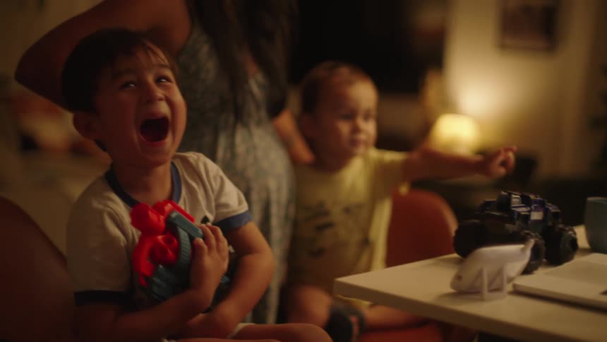 A laptop set on a table with two boys watching it one laughing and the other pointing while holding his mother
