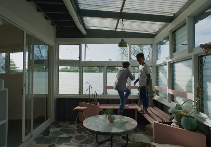 A boy standing on a bench while his brother steps up onto the same bench in a sun room