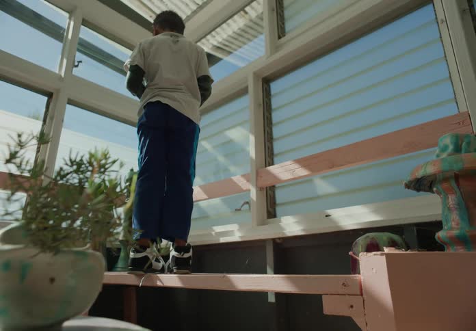 A young boy climbs on top of a wooden desk to look out glass windows in the corner of a room