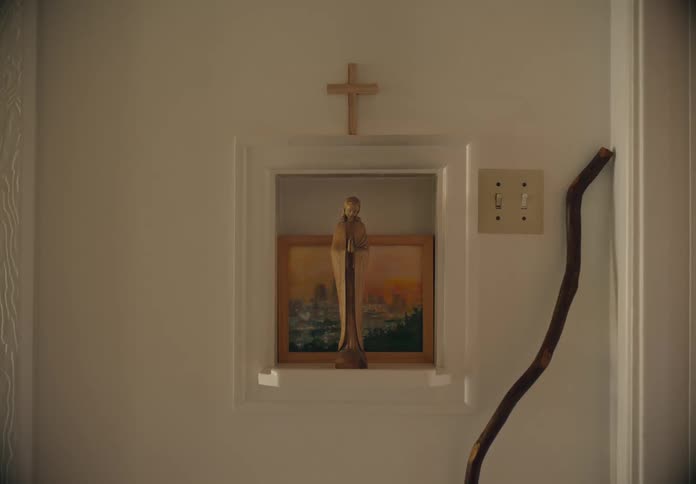 A framed picture with a religious statue in front of it with a cross above it and a handle leaned up against the wall near the light switch