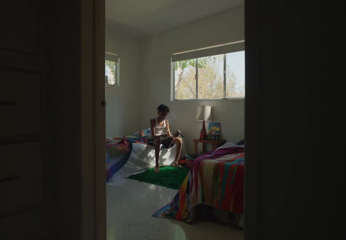 A boy sits on a bed under a window and lights a match